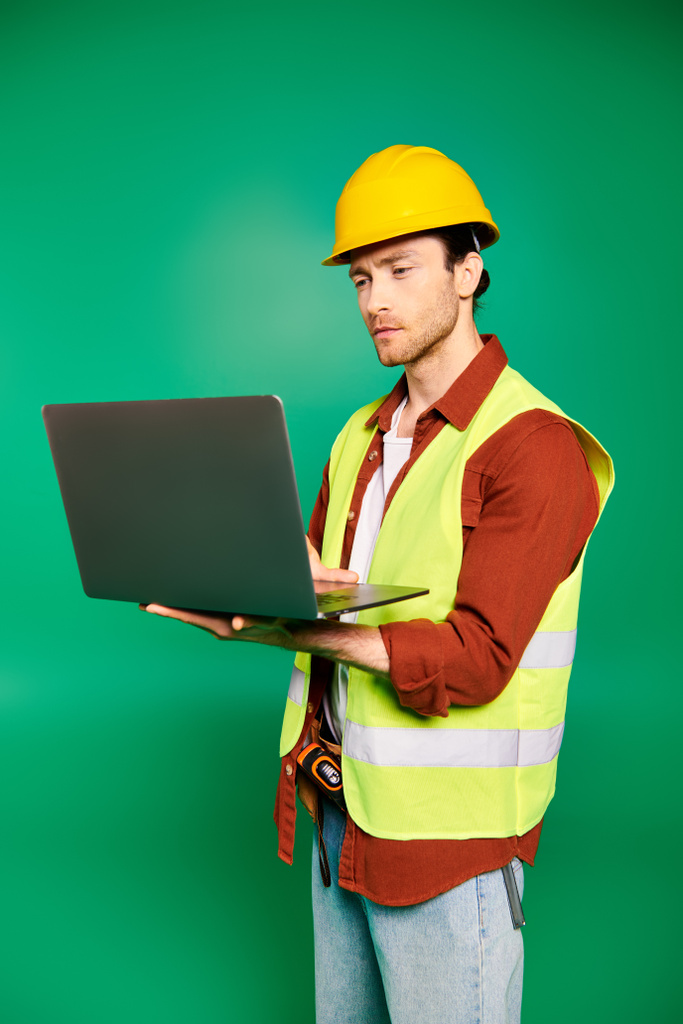 Όμορφος εργάτης με σκληρό καπέλο κρατώντας ένα φορητό υπολογιστή σε ένα πράσινο φόντο. - Φωτογραφία, εικόνα