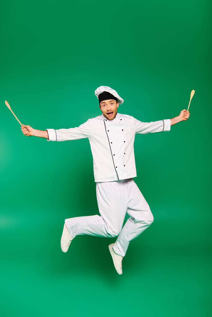 Guapo chef masculino en uniforme blanco saltando alegremente sobre fondo verde. - Foto, Imagen