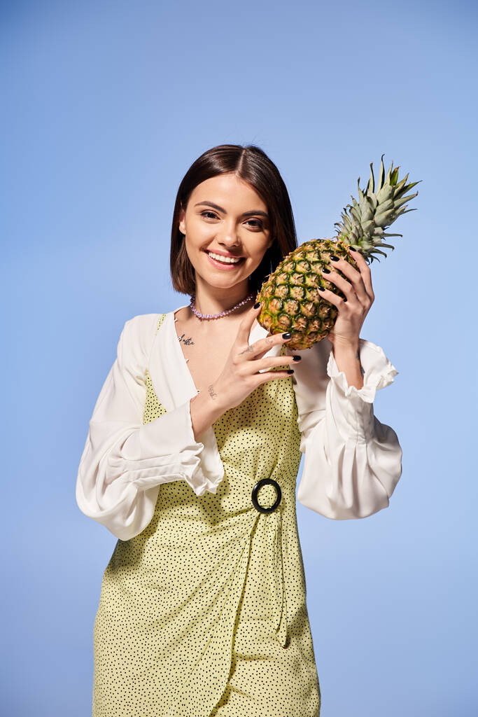 Une jeune femme aux cheveux bruns tient ludique un ananas jusqu'au visage dans un décor de studio. - Photo, image