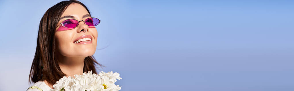 Una giovane donna elegante con capelli castani che indossa occhiali da sole e tiene un vibrante mazzo di fiori in un ambiente da studio. - Foto, immagini