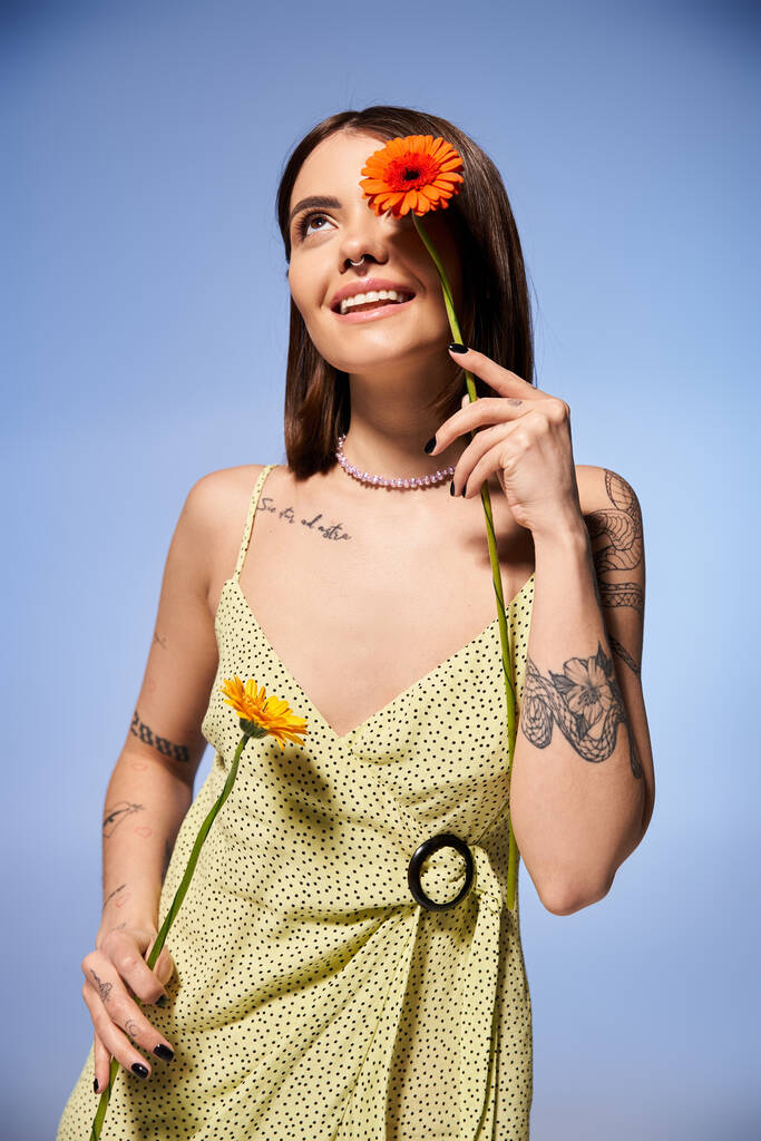 A serene young woman with brunette hair in a bright yellow dress gracefully holds a delicate flower. - Photo, Image