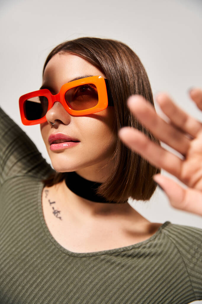 Eine stylische junge Frau mit brünetten Haaren, orangefarbener Sonnenbrille und grünem Hemd im Studio. - Foto, Bild