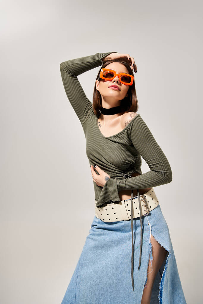 Een brunette vrouw draagt een rok en een zonnebril poseert zelfverzekerd voor een foto in een studio instelling. - Foto, afbeelding