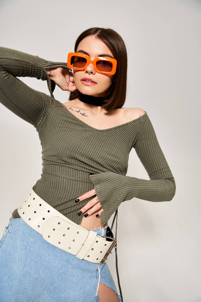 Eine stylische junge Frau mit brünetten Haaren trägt ein grünes Oberteil und eine orangefarbene Sonnenbrille in einem Studio-Ambiente. - Foto, Bild