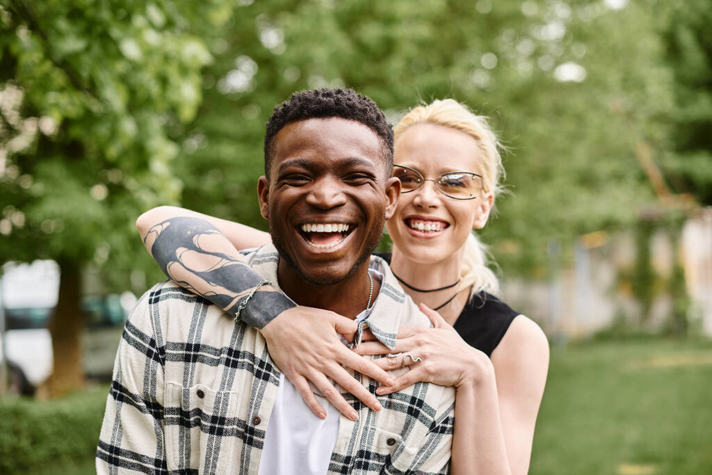 Un hombre afroamericano alegre sostiene a una mujer caucásica en sus brazos, compartiendo un momento de amor al aire libre en un parque. - Foto, imagen