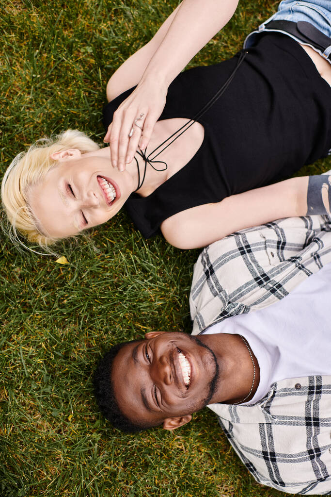 A joyful African American man and Caucasian woman lying together on the green grass, connecting in the peaceful outdoor setting. - Photo, Image