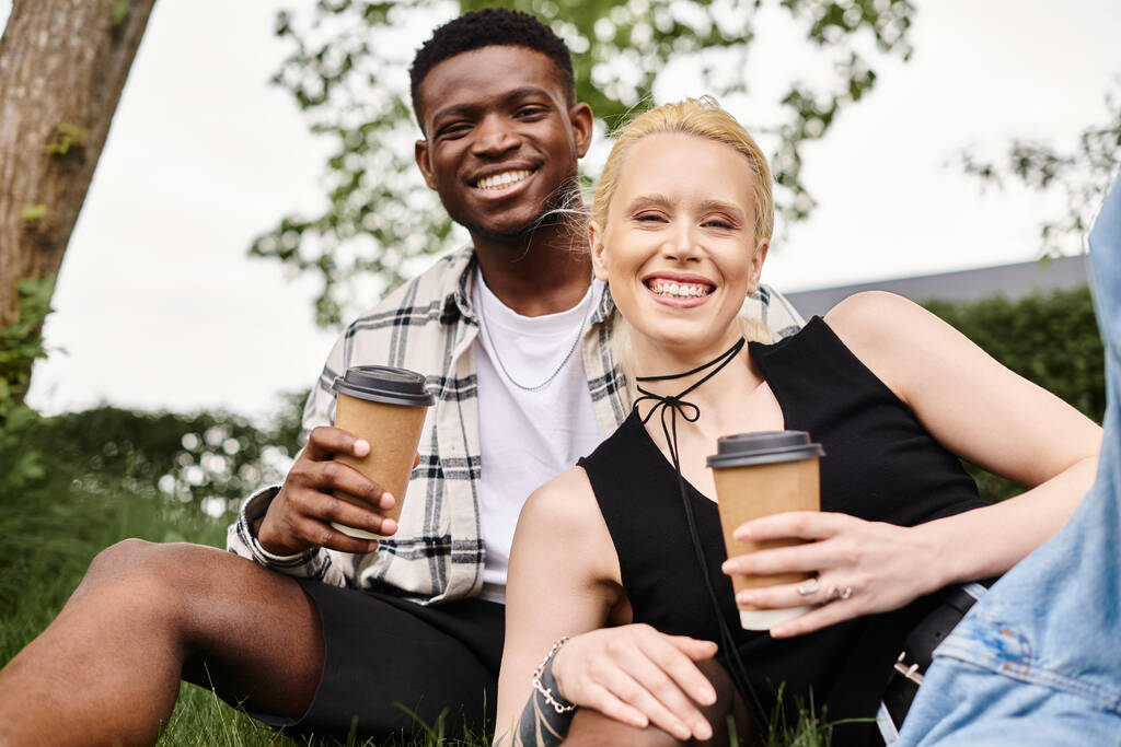 Een gelukkig multicultureel koppel, een Afro-Amerikaanse man en een blanke vrouw, zittend op gras met koffiekopjes. - Foto, afbeelding