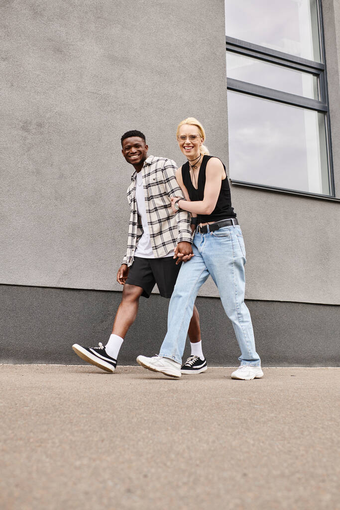 Un heureux petit ami multiculturel et sa petite amie marchent ensemble dans une rue urbaine près d'un bâtiment gris. - Photo, image