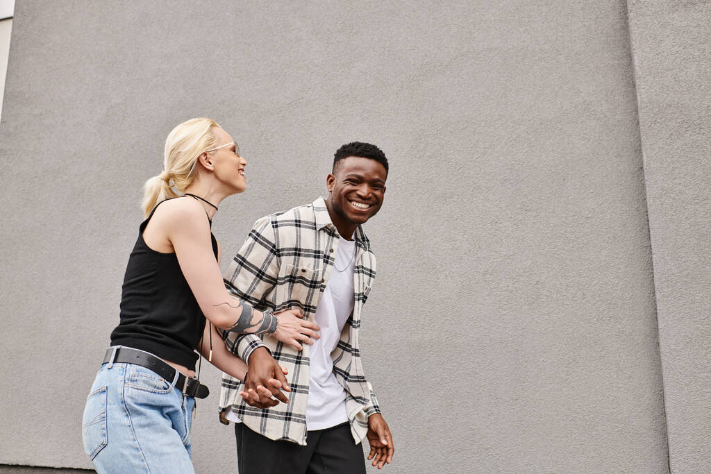 Ένα ευτυχισμένο ζευγάρι, ένας πολυπολιτισμικός φίλος και η φίλη του, περπατούν μαζί σε έναν αστικό δρόμο κοντά σε ένα γκρίζο κτίριο. - Φωτογραφία, εικόνα