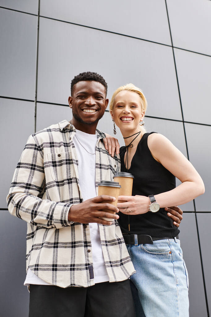Una pareja multicultural, un hombre y una mujer, están felices uno al lado del otro en una calle urbana cerca de un edificio gris. - Foto, Imagen