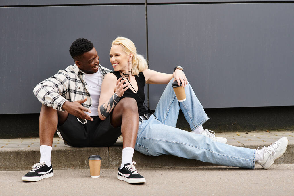Een man en een vrouw van verschillende etniciteiten kijken tevreden als ze naast elkaar zitten op de grond in de buurt van een grijs gebouw in de stad. - Foto, afbeelding