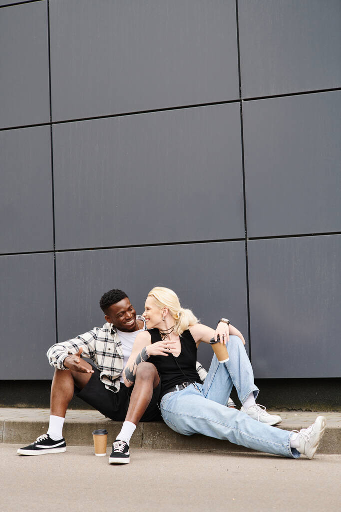 Una feliz pareja multicultural sentada una al lado de la otra en el suelo cerca de un edificio gris urbano, compartiendo un momento de tranquilidad. - Foto, imagen
