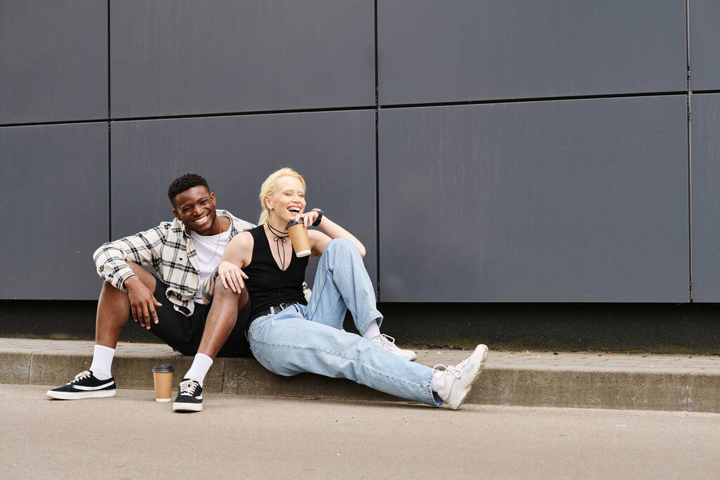 Ein fröhliches multikulturelles Paar sitzt nebeneinander auf dem Boden in der Nähe eines grauen städtischen Gebäudes - Foto, Bild