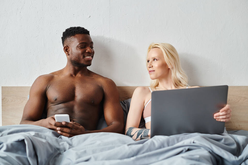 Ένας πολυπολιτισμικός άνδρας και μια γυναίκα κάθονται κοντά σε ένα κρεβάτι, απορροφημένοι στην οθόνη ενός φορητού υπολογιστή μπροστά τους. - Φωτογραφία, εικόνα