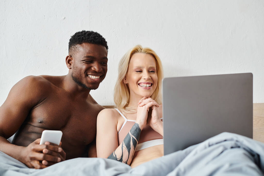Ένα πολυπολιτισμικό ζευγάρι κάθεται σε ένα κρεβάτι, απορροφημένο σε μια οθόνη laptop. - Φωτογραφία, εικόνα
