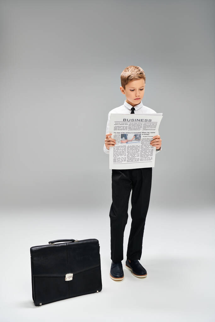 Niño preescolar vestido con elegante atuendo de pie junto a una maleta. Concepto de negocio sobre fondo gris. - Foto, Imagen