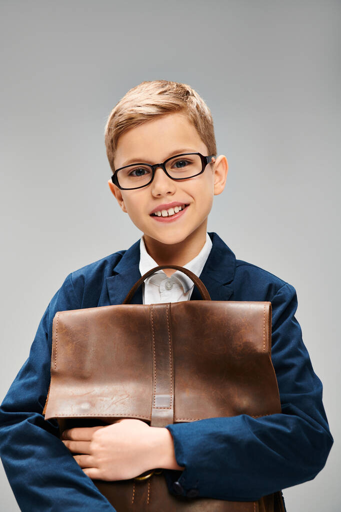 Een jonge jongen in elegante kleding, draagt een bril, houdt een bruine tas op een grijze achtergrond. - Foto, afbeelding
