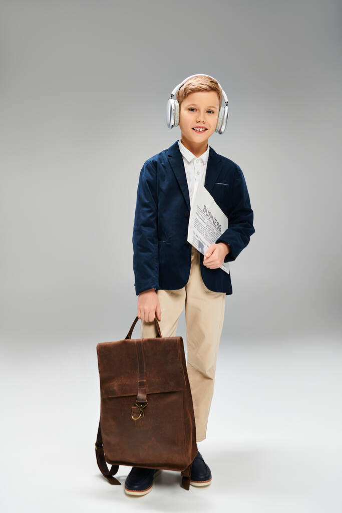 Предрожденный мальчик в элегантной одежде, в наушниках, держит портфель на сером фоне. - Фото, изображение