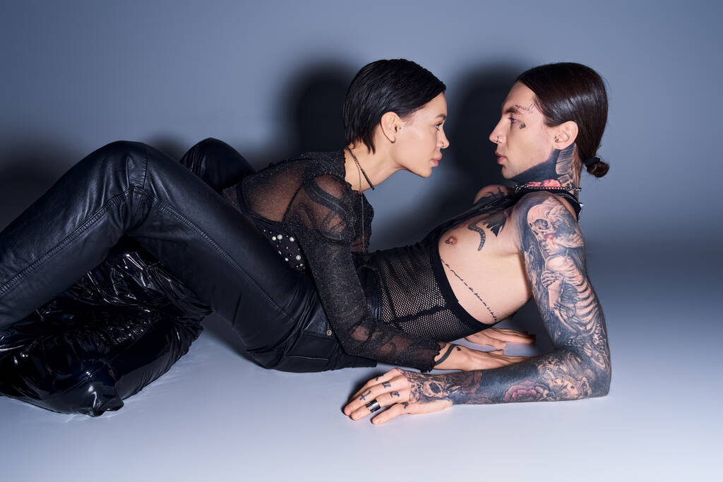 Um jovem casal tatuado compartilha um momento próximo contra um cenário cinza sereno. - Foto, Imagem