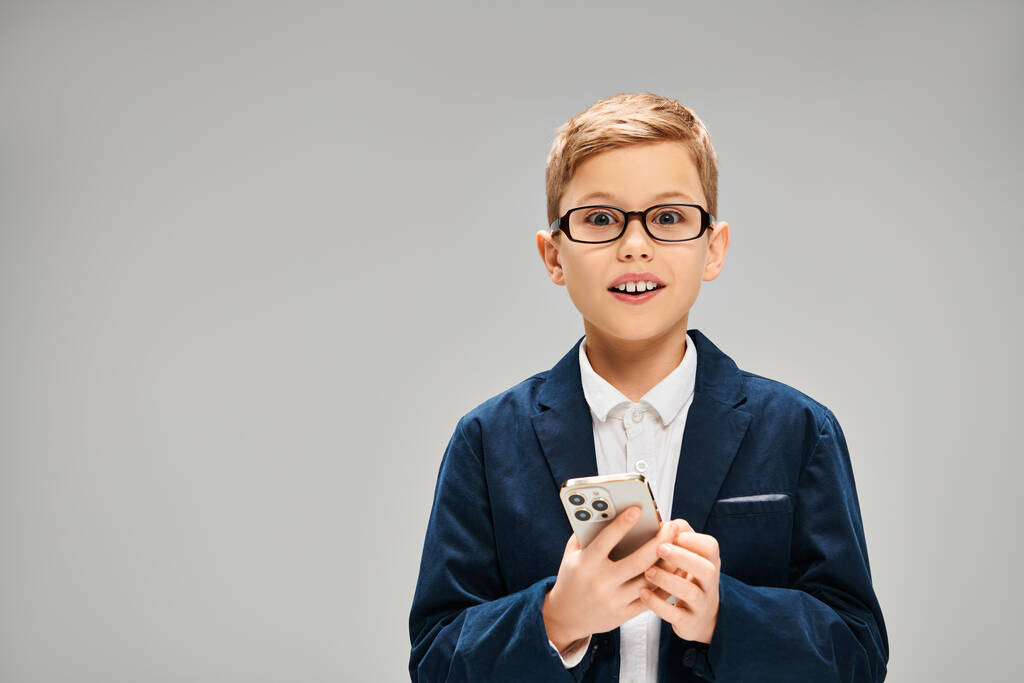 Elegante niño preadolescente en gafas con teléfono celular sobre fondo gris. - Foto, imagen