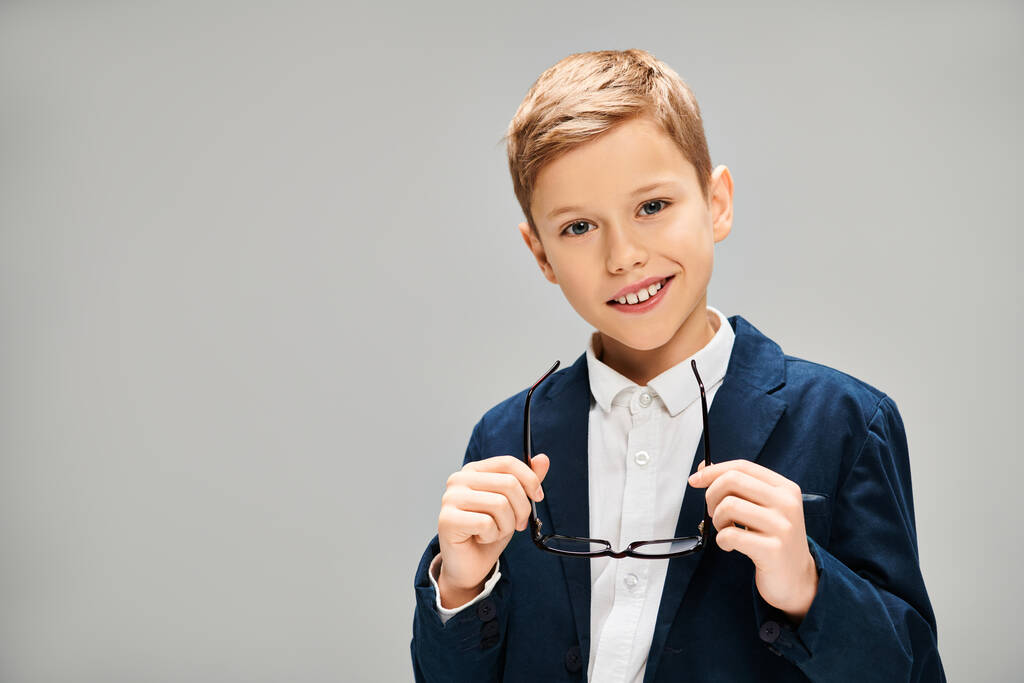 Niño en traje elegante sostiene un par de gafas contra un telón de fondo gris. - Foto, imagen