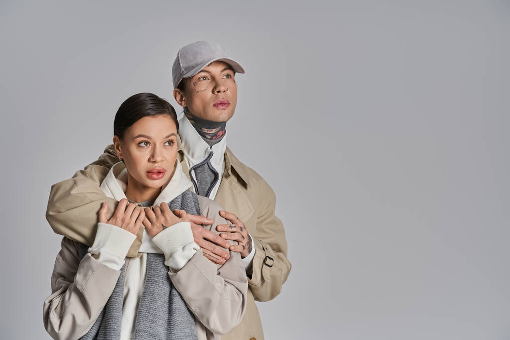 トレンチコートを着用したスタイリッシュな若い男性と女性がスタジオの横に立って灰色の背景. - 写真・画像