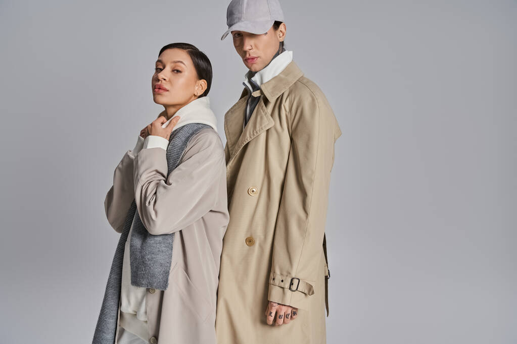 Um jovem casal elegante em casacos de trincheira de pé ao lado um do outro em um estúdio contra um fundo cinza. - Foto, Imagem