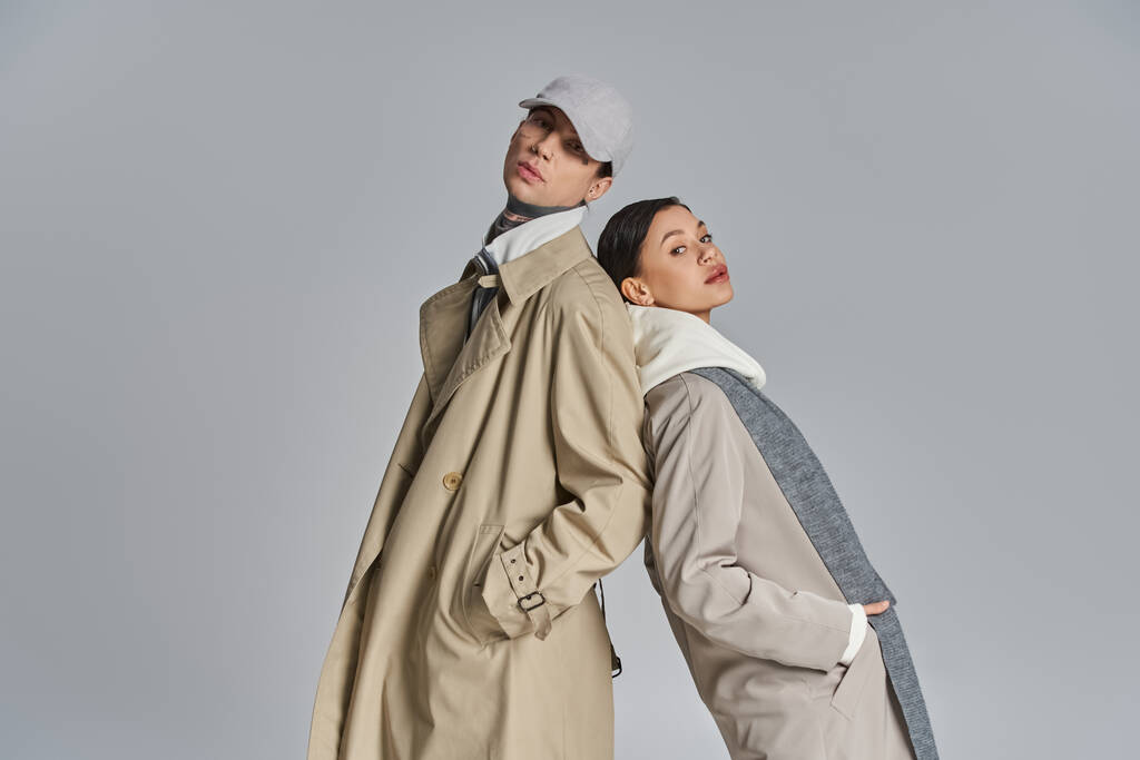 Un jeune homme et une jeune femme debout côte à côte en trench coats, exsudant style urbain et élégance sur fond de studio gris. - Photo, image
