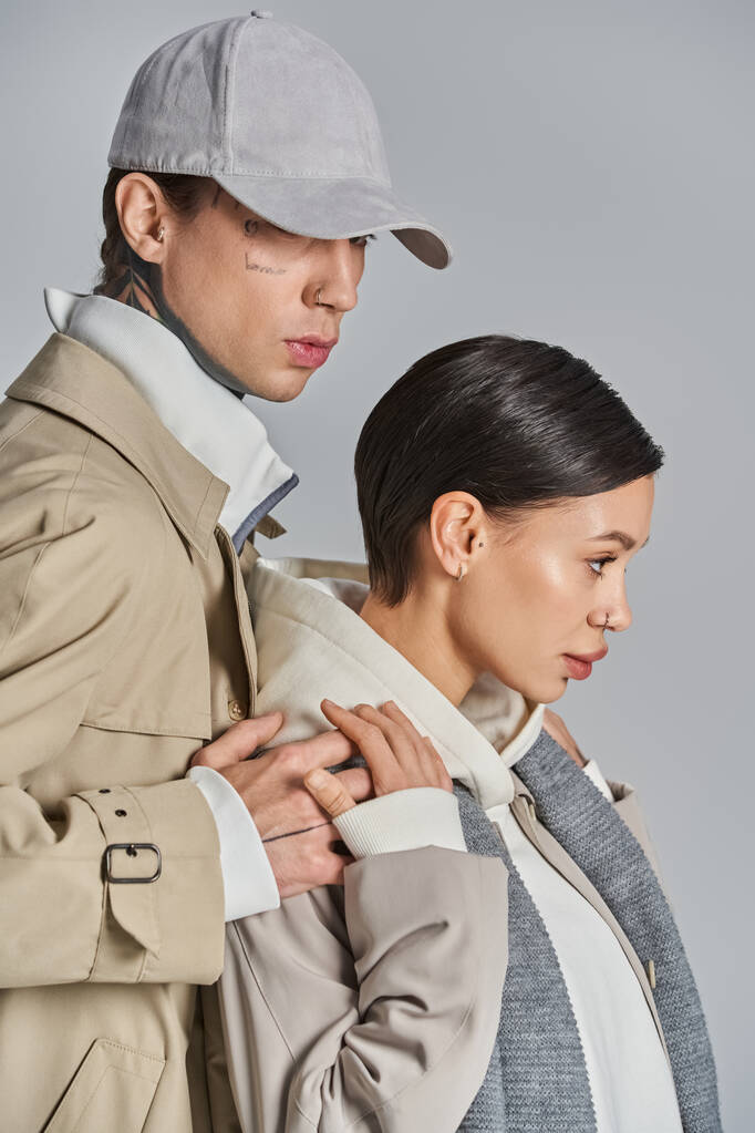 Una pareja joven y elegante está de pie lado a lado en abrigos de trinchera en un estudio sobre un fondo gris. - Foto, imagen