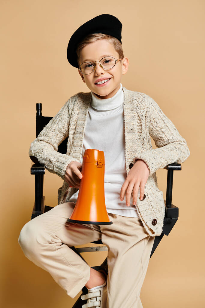 Мальчик, одетый как кинорежиссёр, держит в руках оранжевый мегафон, сидя в кресле. - Фото, изображение