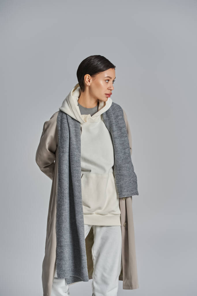 Uma jovem mulher elegante fica confiante em um casaco de trincheira contra um fundo cinza liso. - Foto, Imagem