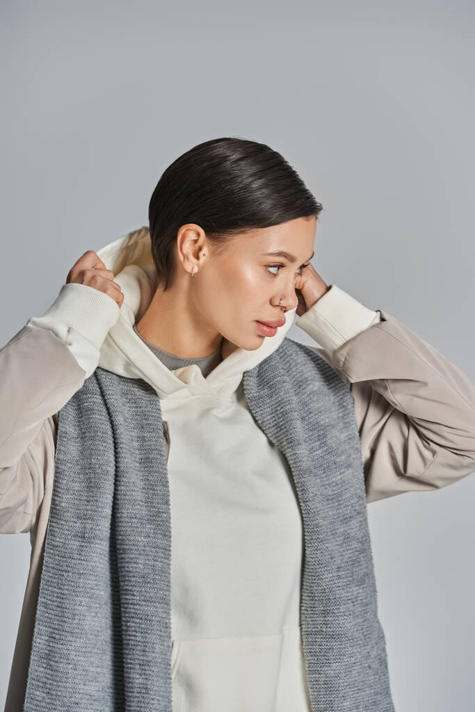 Eine junge Frau in einem weißen Oberteil drapiert anmutig einen grauen Schal und strahlt in einem Atelier vor grauem Hintergrund Raffinesse aus.. - Foto, Bild