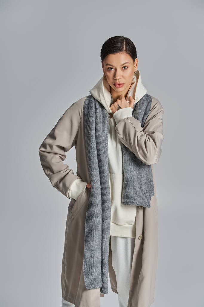 A stylish young woman exudes elegance in a gray trench coat and white pants against a neutral studio backdrop. - Photo, Image