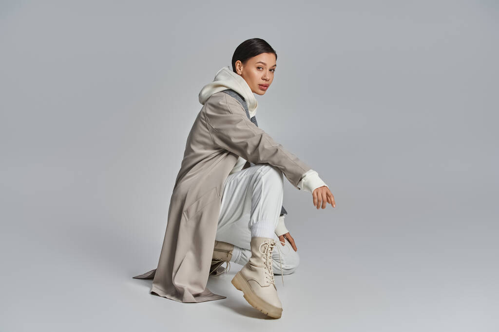 A young stylish woman in a trench coat sitting gracefully in a studio against a grey background. - Photo, Image