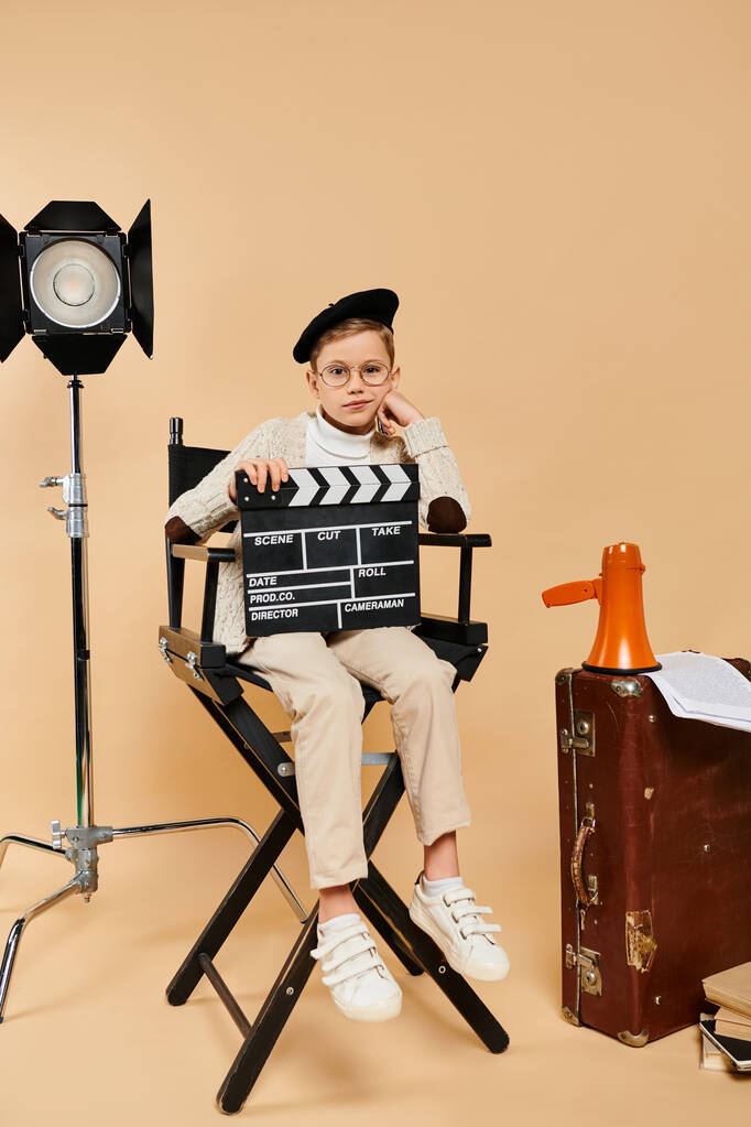 Předdospívající chlapec v režisérském oděvu s filmovým tleskačem, sedící v křesle. - Fotografie, Obrázek