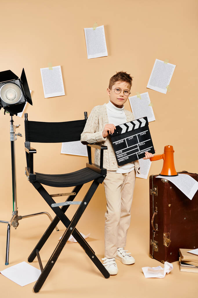 椅子の隣に映画のクラッパーを保持する映画監督の衣装の若い少年. - 写真・画像