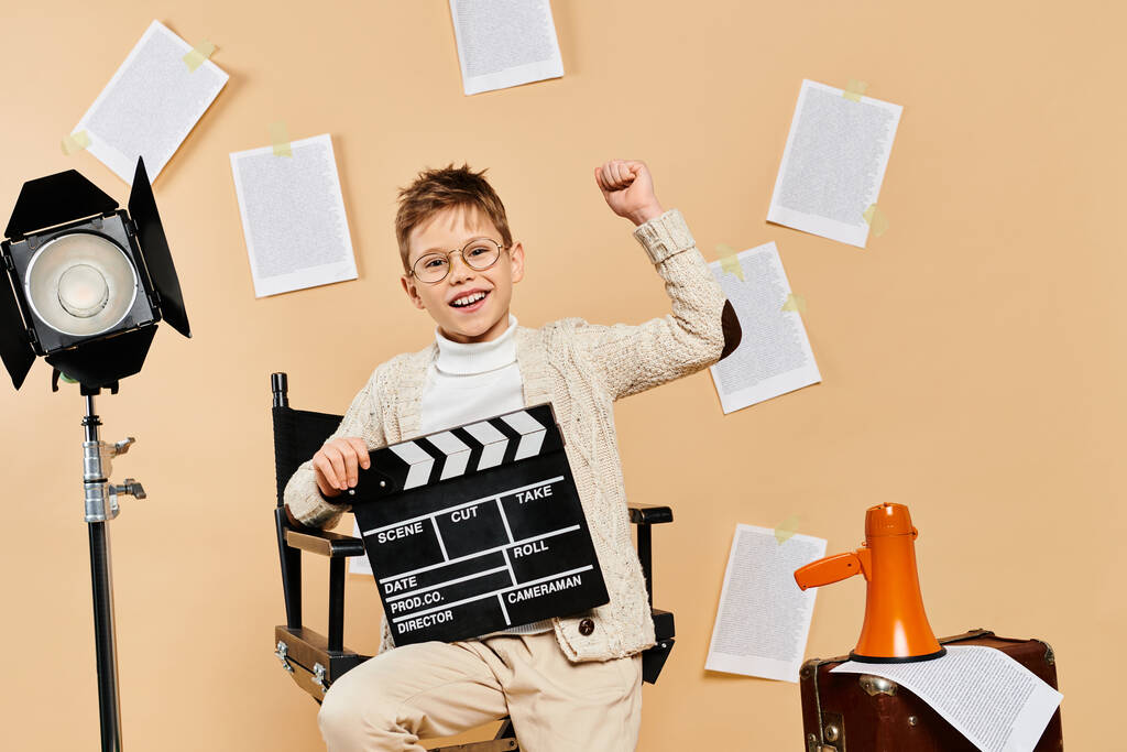 Мальчик-подросток, одетый как кинорежиссёр, сидит на бежевом фоне с аплодисментами.. - Фото, изображение