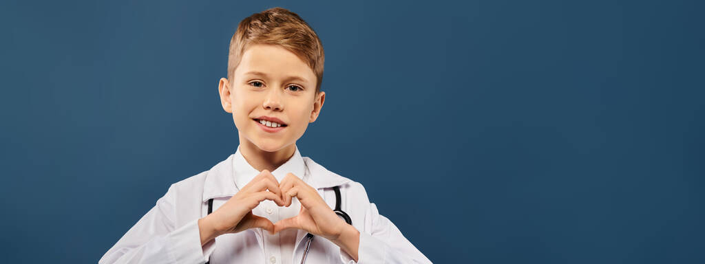 Nuori poika pukeutunut lääkäri muodostaa sydämen muodon kädet sinisellä taustalla. - Valokuva, kuva