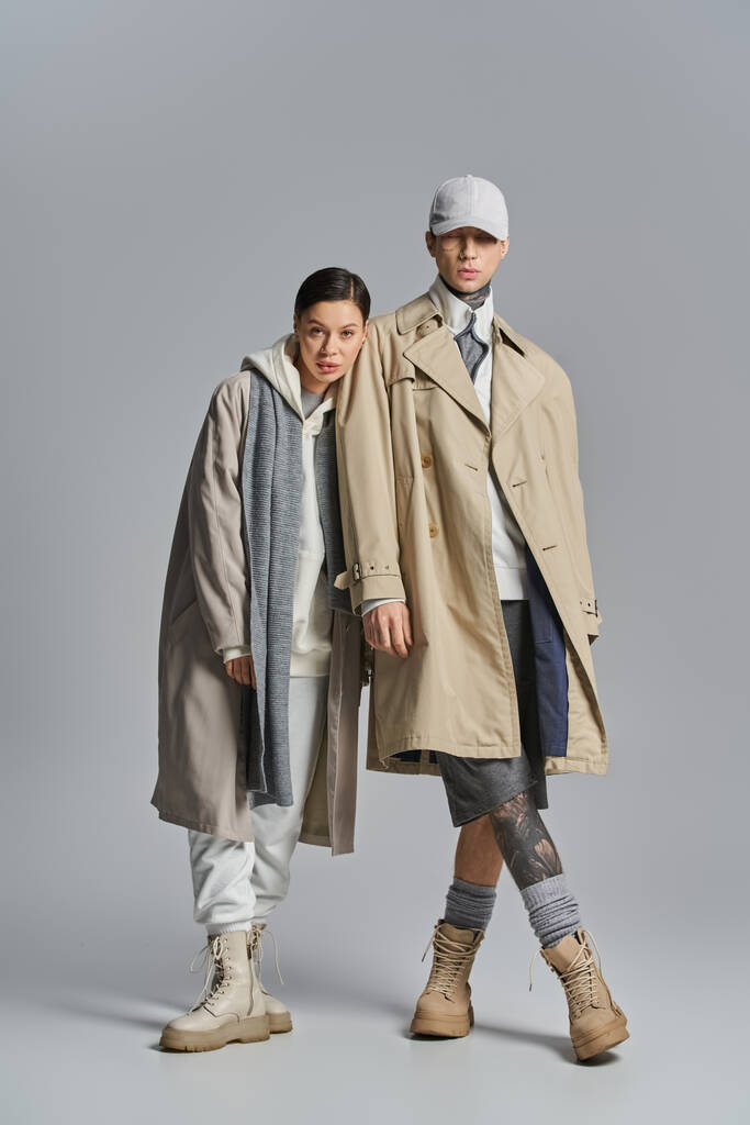 Ein junges, stylisches Paar steht nebeneinander in Trenchcoats in einem Atelier vor grauem Hintergrund. - Foto, Bild