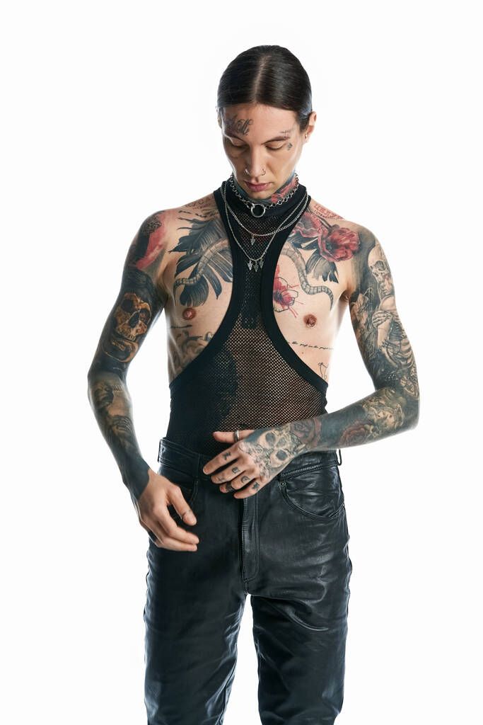 Młody mężczyzna z uderzającymi tatuażami i kolczykami na klatce piersiowej pozuje w studio na szarym tle.. - Zdjęcie, obraz