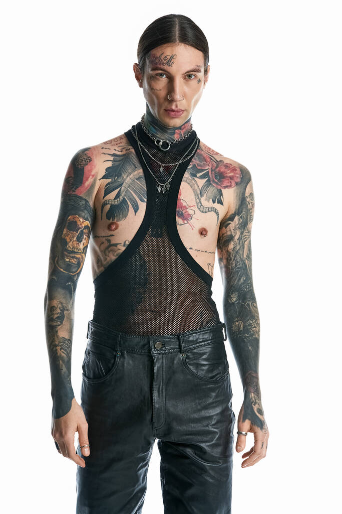Ένας νεαρός άνδρας με πολλά τατουάζ που κοσμούν το σώμα του ποζάρει με αυτοπεποίθηση σε ένα στούντιο πάνω σε γκρι φόντο.. - Φωτογραφία, εικόνα