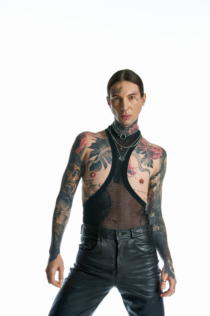Ένας άντρας με τατουάζ αποπνέει στάση με δερμάτινα παντελόνια, δείχνοντας το τολμηρό στυλ του σε ένα γκρίζο σκηνικό στούντιο. - Φωτογραφία, εικόνα