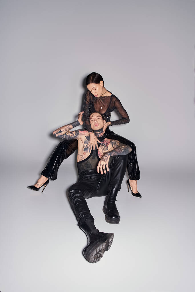 Молодая стильная женщина сидит на вершине мужчины в студии, оба покрыты татуировками, на сером фоне. - Фото, изображение