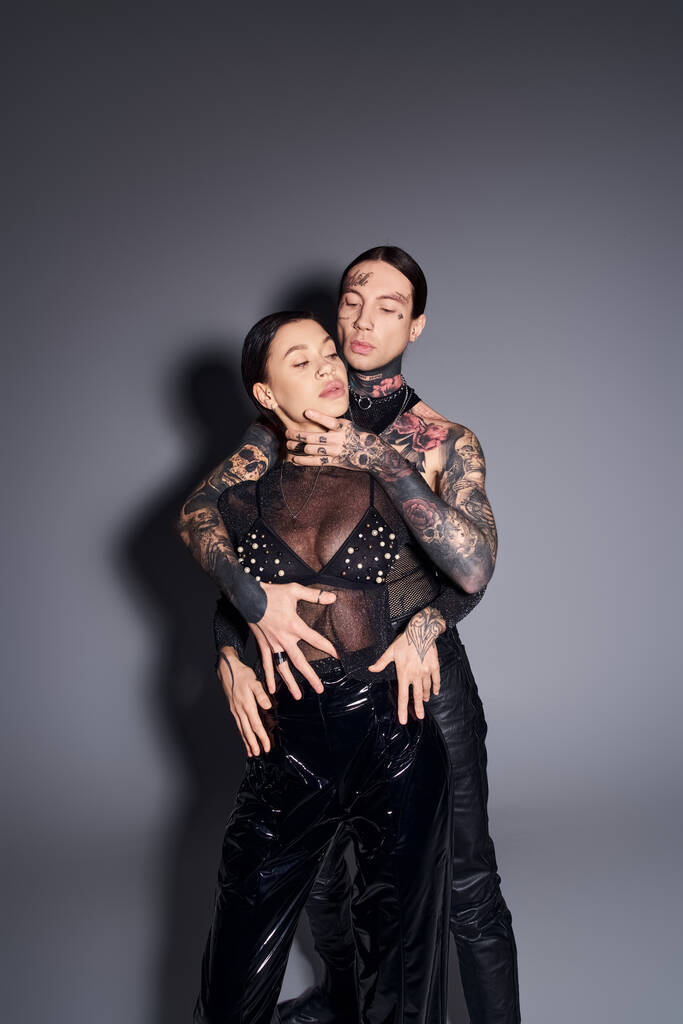 Una pareja joven y elegante con tatuajes vestidos con trajes de cuero negro a juego posan en un estudio sobre un fondo gris. - Foto, imagen