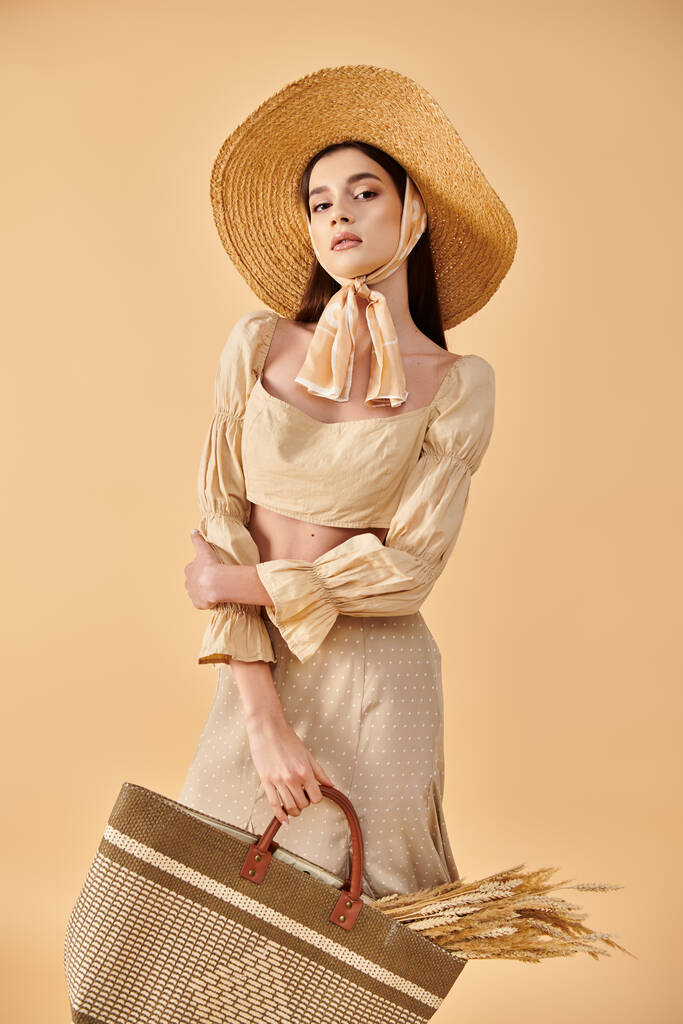 Κομψό νεαρή γυναίκα με μακριά καστανά μαλλιά σε ένα καπέλο και φόρεμα κρατώντας ένα καλάθι, ενσωματώνοντας μια καλοκαιρινή αύρα σε ένα σκηνικό στούντιο. - Φωτογραφία, εικόνα