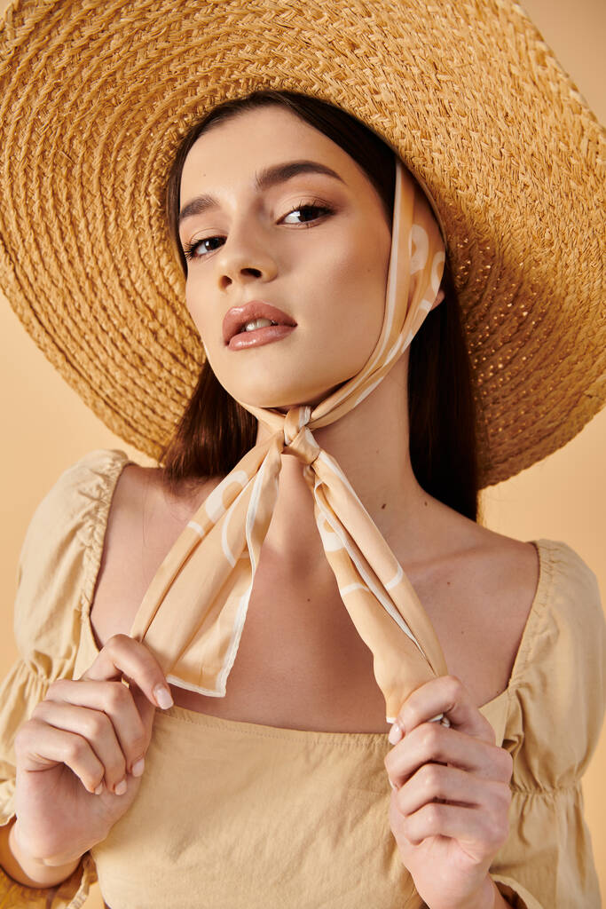 Молодая женщина с длинными брюнетками позирует в летнем наряде, в соломенной шляпе и шарфе, излучая безмятежную и загорелую атмосферу. - Фото, изображение