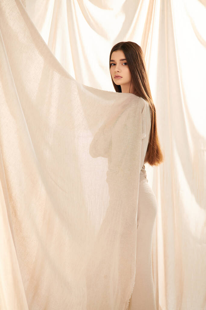 Una giovane donna con lunghi capelli castani posa davanti a una tenda bianca, trasudando un umore estivo nel suo vestito elegante. - Foto, immagini