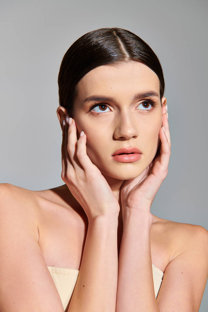 A young woman with brunette hair poses in a studio setting with her hands delicately placed on her face. - Photo, Image