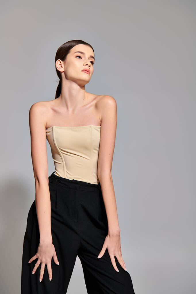 A stylish young woman with brunette hair strikes a pose in a studio setting, showcasing a trendy tan top paired with classic black pants. - Photo, Image