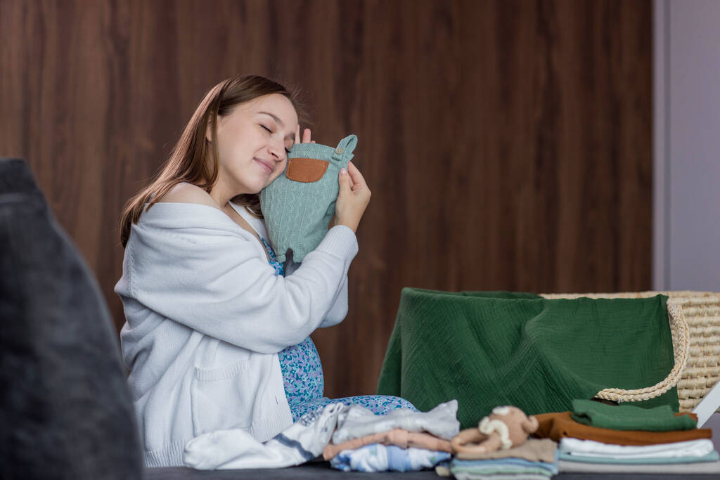 Mulher grávida dobrar roupas de bebê sentado no sofá em casa. - Foto, Imagem